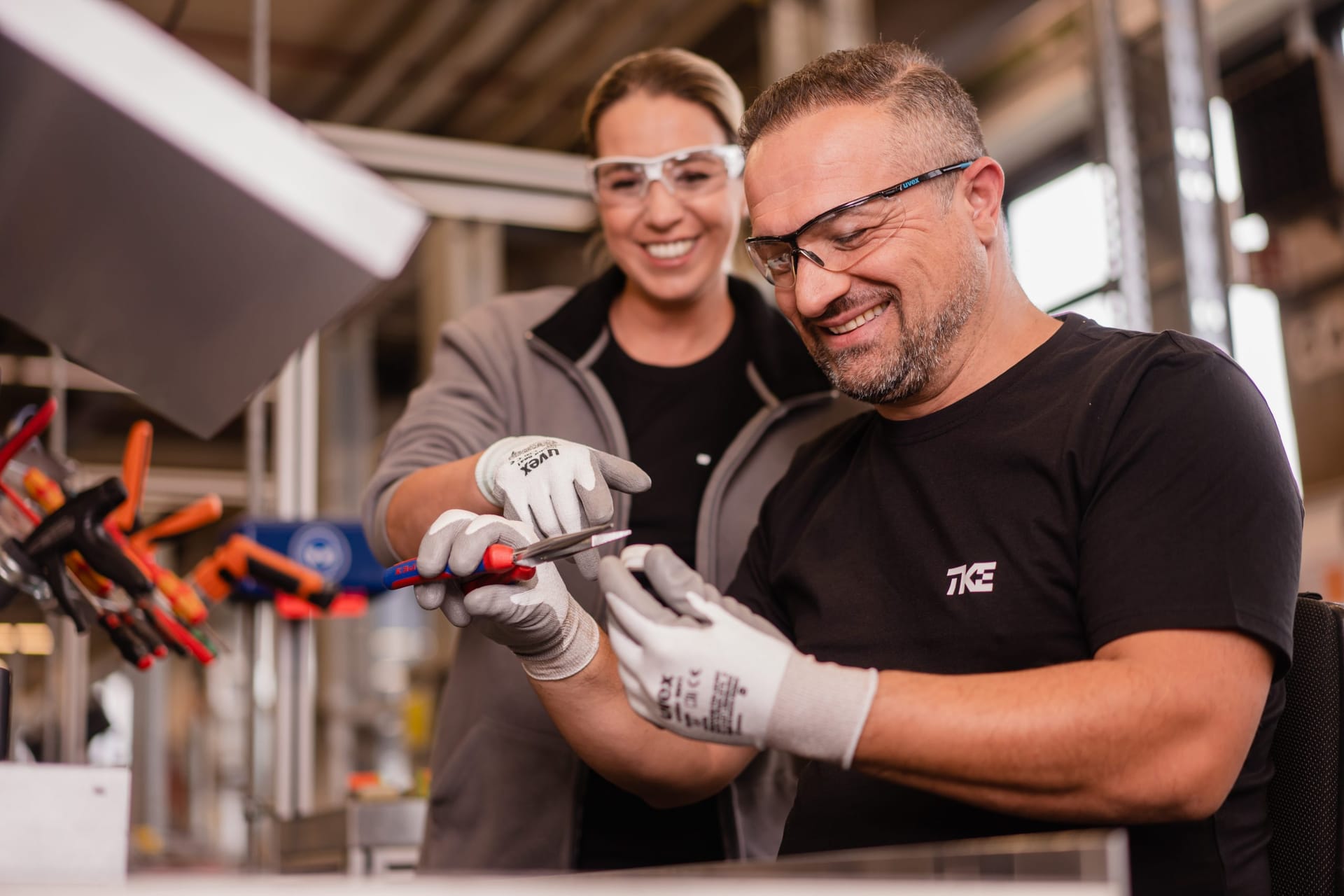 Dois colegas sorridentes com óculos de proteção e luvas de trabalho manuseiam juntos uma ferramenta em uma bancada.