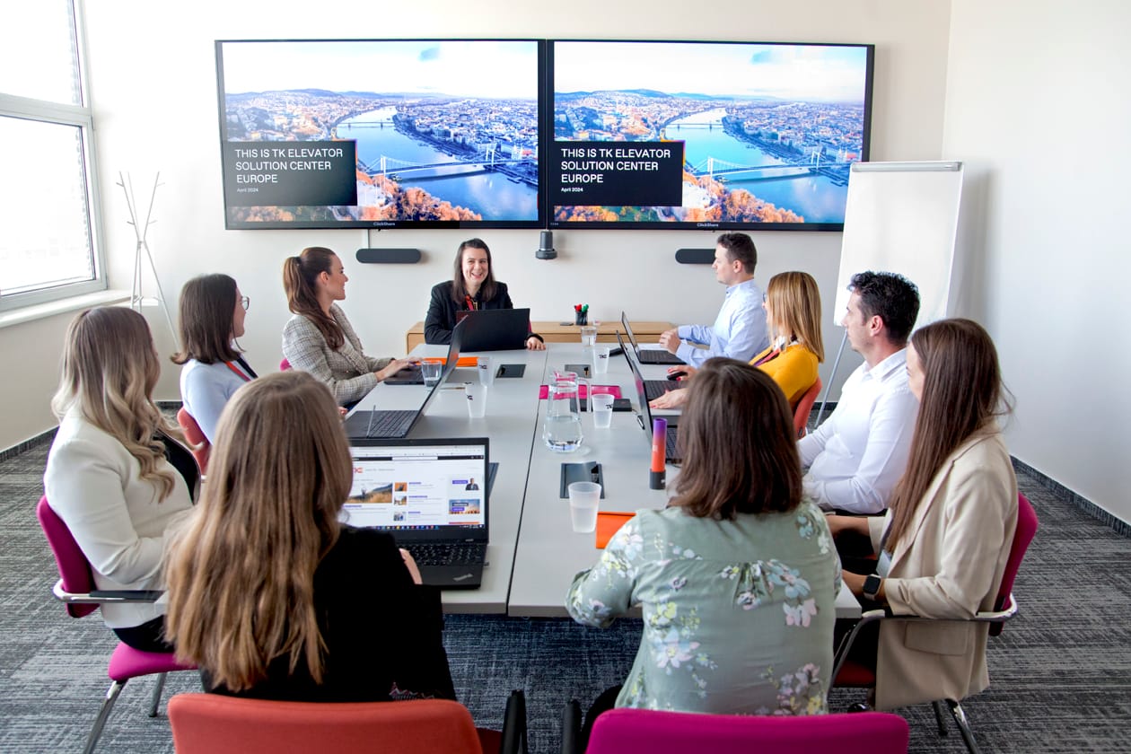 Uma equipe se reúne em torno de uma longa mesa de conferência sob duas telas com o título “This is TK Elevator Solution Center Europe”.