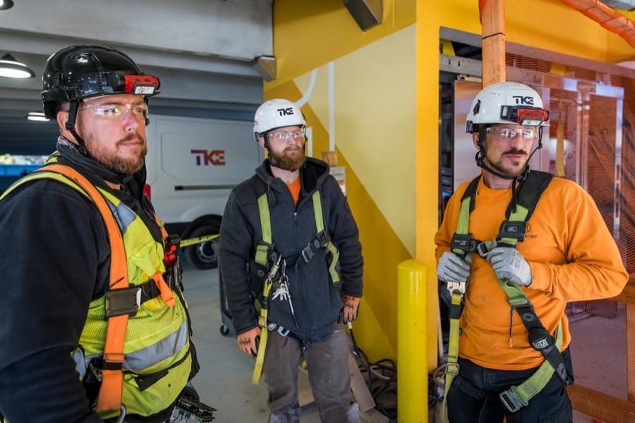 Três técnicos da TKE com capacete, óculos de proteção e cinto de segurança em uma obra.